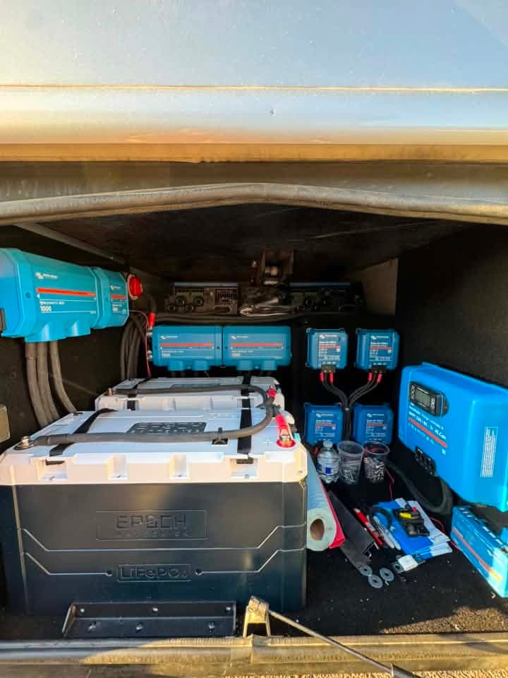 Two Epoch LiFePO4 batteries strapped into a carpeted RV storage bay beside Victron Lynx modules, chargers and a solar charge controller